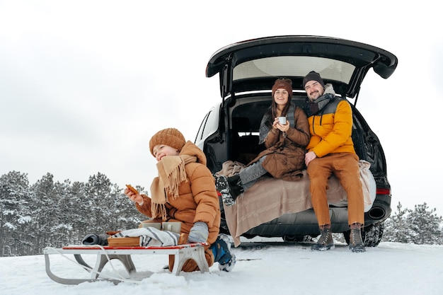 Portrait d'hiver d'une famille assise sur le coffre d'une voiture profitant de leurs vacances en forêt