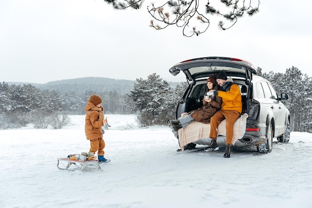 Portrait d'hiver d'une famille assise sur le coffre d'une voiture profitant de leurs vacances en forêt
