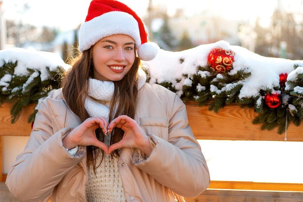 Portrait d'hiver jeune adulte belle femme en bonnet de Noel montrant la forme de coeur par les doigts Ambiance de Noël Concept de mode de beauté d'hiver de neige