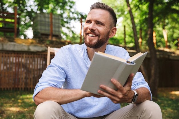 Portrait d'un homme adulte heureux assis dans une chaise longue et livre de lecture, pendant le repos dans le parc verdoyant