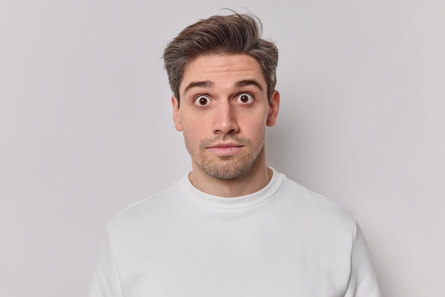 Portrait d'un homme adulte surpris et choqué regarde les yeux obsédés par la caméra et entend de grandes nouvelles vêtus d'un pull décontracté isolé sur fond blanc. Le jeune homme concerné se sent très surpris pose à l'intérieur