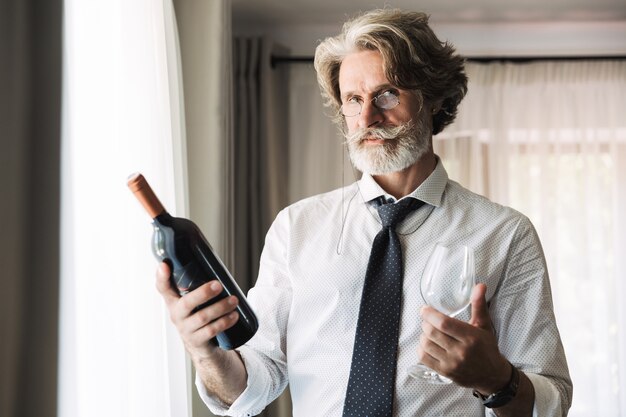 Portrait d'homme d'affaires adulte barbu à lunettes vêtus de vêtements de cérémonie tenant une bouteille de vin et de verre dans un appartement de l'hôtel