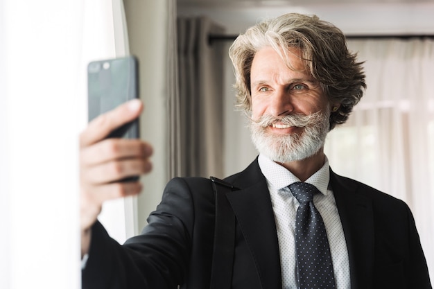 Portrait d'un homme d'affaires adulte heureux portant un costume noir prenant un selfie sur un smartphone tout en se tenant près de la fenêtre dans un appartement de l'hôtel