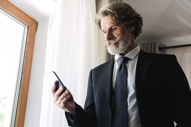 Portrait d'un homme d'affaires adulte portant un costume noir tenant un smartphone tout en se tenant près de la fenêtre dans un appartement de l'hôtel