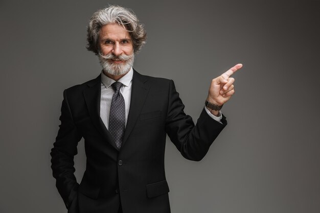 Portrait d'un homme d'affaires adulte prospère portant un costume noir formel souriant et pointant du doigt sur un fond isolé sur un mur gris