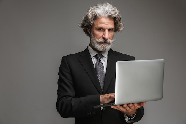 Photo portrait d'un homme d'affaires adulte sérieux portant un costume noir formel tenant un ordinateur portable argenté isolé sur un mur gris