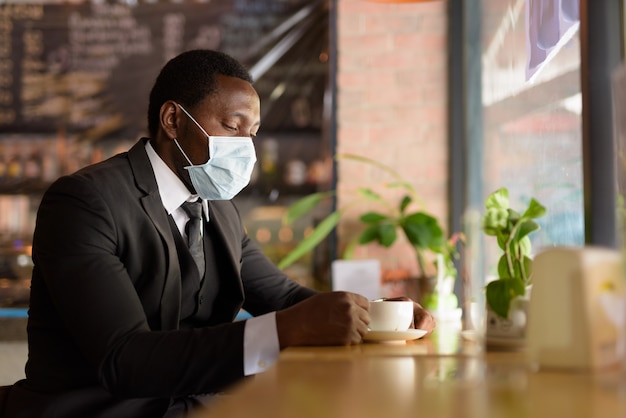 Portrait d'homme d'affaires africain portant un masque tout en buvant du café à l'intérieur du café comme nouvelle norme