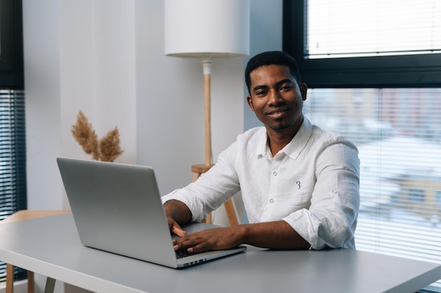 Portrait d'un homme d'affaires afro-américain positif utilisant un ordinateur portable et souriant regardant la caméra tout en appréciant le travail assis au bureau dans une salle de bureau moderne