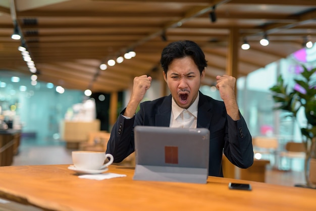 Portrait d'homme d'affaires asiatique dans un café avec les bras levés à l'aide d'une tablette numérique