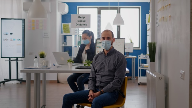 Portrait d'un homme d'affaires assis au bureau portant un masque médical contre l'infection par le coronavirus pendant la pandémie mondiale. Les travailleurs d'équipe travaillant sur un projet d'entreprise maintiennent une distance sociale