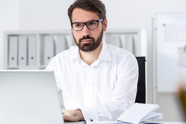 Portrait d'un homme d'affaires barbu assis devant son ordinateur portable dans un bureau blanc et pensant en regardant au loin. Concept de planification