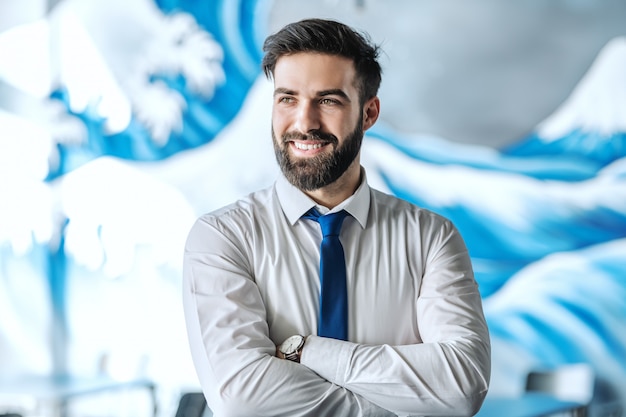 Portrait d'homme d'affaires beau sourire en chemise et cravate debout dans le bureau avec les bras croisés et en détournant les yeux.