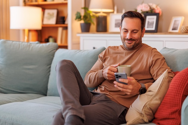 Portrait d'un homme d'affaires confiant tenant un café et envoyant des SMS sur son téléphone portable alors qu'il est assis sur le canapé à la maison