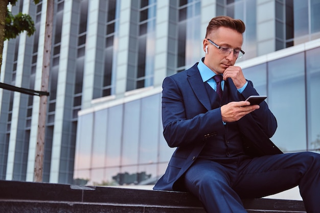 Portrait d'un homme d'affaires élégant et confiant vêtu d'un costume élégant à l'aide d'un smartphone assis à l'extérieur sur un fond de gratte-ciel.