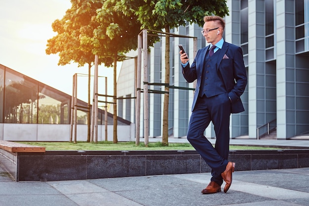 Photo portrait d'un homme d'affaires élégant et confiant vêtu d'un costume élégant à l'aide d'un smartphone tout en se tenant à l'extérieur sur un fond de gratte-ciel.