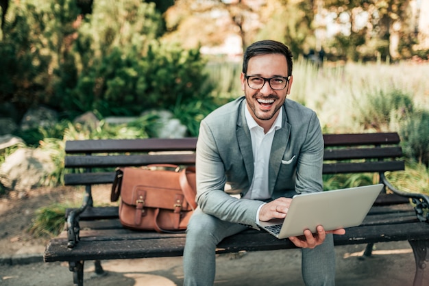 Portrait d&#39;un homme d&#39;affaires heureux à l&#39;aide d&#39;un ordinateur portable à l&#39;extérieur. Assis sur le banc et regardant la caméra.