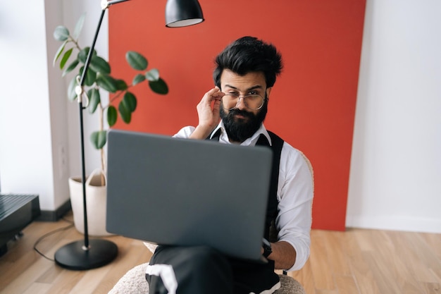 Portrait d'un homme d'affaires indien positif en tenues de soirée travaillant sur un ordinateur portable assis sur une chaise dans un bureau de coworking léger regardant la caméra