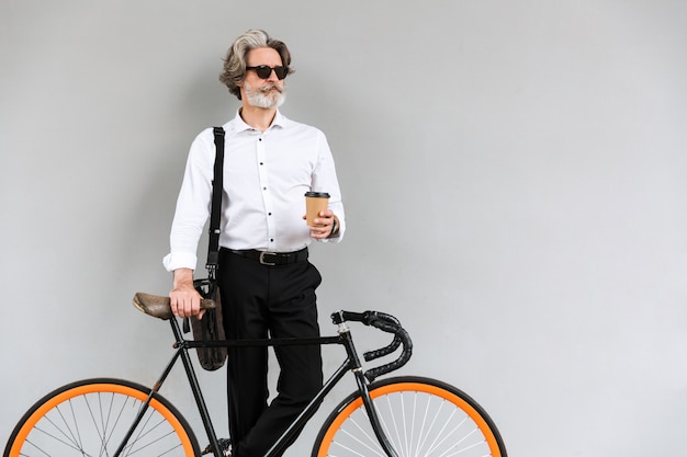 Portrait d'homme d'affaires mature masculin dans des lunettes de soleil buvant du café à emporter en se tenant debout avec un vélo sur un mur gris à l'extérieur