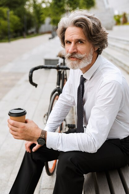 Portrait d'homme d'affaires mûr sérieux dans des vêtements formels buvant du café à emporter tout en étant assis sur un banc avec un vélo à l'extérieur