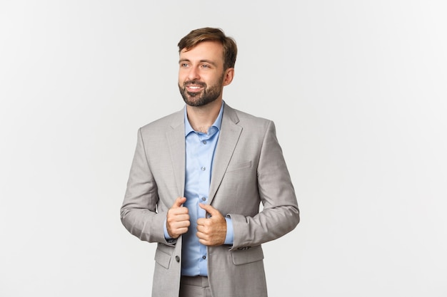 Portrait d'homme d'affaires prospère et confiant en costume gris et chemise bleue, souriant heureux et regardant à gauche, debout sur fond blanc.