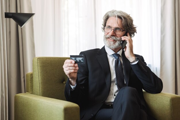 Portrait d'un homme d'affaires senior souriant et joyeux aux cheveux gris à l'intérieur à la maison vêtu de vêtements formels tenant une carte de crédit parlant par téléphone portable.