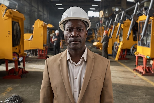 Portrait d'un homme d'affaires sérieux africain dans un casque de travail regardant la caméra travaillant dans une usine avec une nouvelle machine en arrière-plan