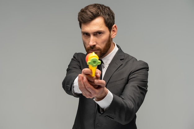 Portrait d'un homme d'affaires sérieux jouant avec un pistolet à eau sur fond gris et regardant la caméra avec un visage calme
