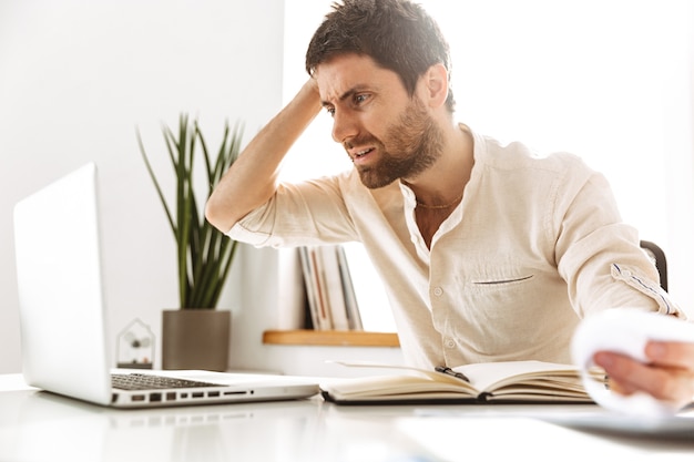 Portrait d'homme d'affaires tendu de 30 ans portant une chemise blanche travaillant avec un ordinateur portable et des documents papier, alors qu'il était assis dans un bureau lumineux