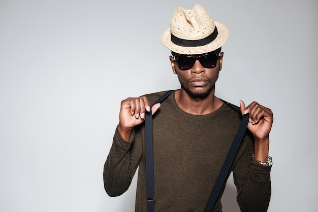 Portrait d'un homme africain portant un chapeau et des lunettes debout en studio. Isolé sur fond gris.