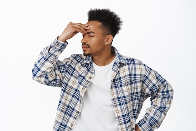 Portrait d'un homme afro-américain agacé qui a l'air fatigué et fatigué, se frotte les yeux et le nez, essayant de se calmer, affligé par une mauvaise situation difficile, debout sur fond blanc