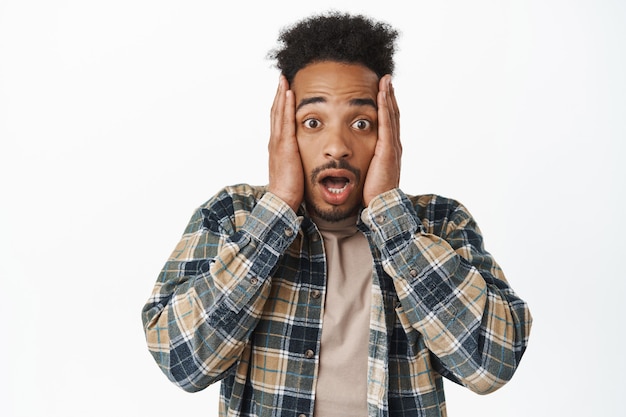 Portrait d'un homme afro-américain choqué haletant sans voix, tenant les mains sur le visage et regardant sans voix avec admiration, vérifiant les grandes nouvelles sur blanc