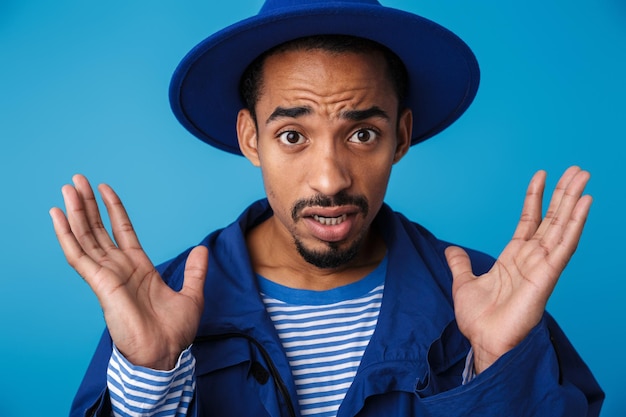 Portrait d'un homme afro-américain confus portant un chapeau avec les mains en l'air isolé sur bleu