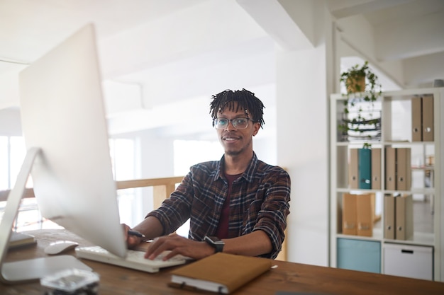 Portrait d'un homme afro-américain contemporain souriant à la caméra tout en utilisant un ordinateur et en tapant sur le clavier au bureau blanc, concept de développeur informatique, espace de copie