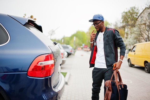 Portrait d'un homme afro-américain élégant sur une casquette de sport et des lunettes marchant avec un sac à main et un coffre de voiture ouvert Hommes noirs modèle mode de rue