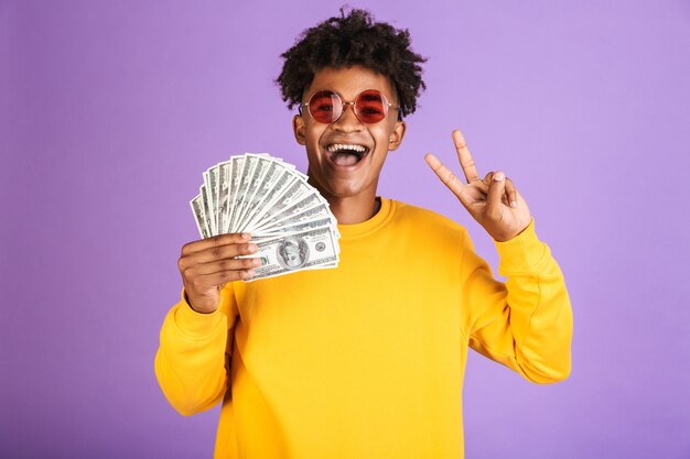 Portrait d'un homme afro-américain élégant portant des lunettes de soleil souriant tout en tenant des billets d'argent en dollars, isolé sur fond violet