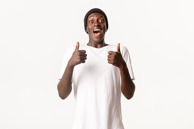 Portrait d'un homme afro-américain excité et joyeux en bonnet, à la recherche amusée et montrant le pouce en l'air en approbation