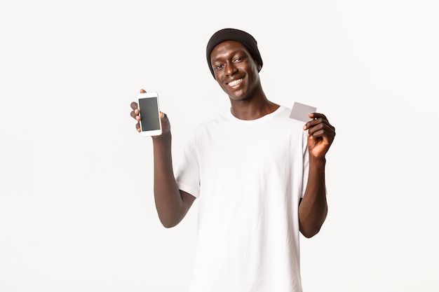 Portrait d'un homme afro-américain heureux et souriant en bonnet, montrant l'écran du smartphone et la carte de crédit