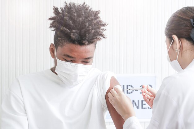 Portrait d'un homme afro-américain regardant tout en se faisant vacciner contre le covid dans une clinique ou un hôpital, avec une infirmière à main s'injectant un vaccin pour obtenir une immunité pour protéger le virus. adolescent portant un masque de protection.