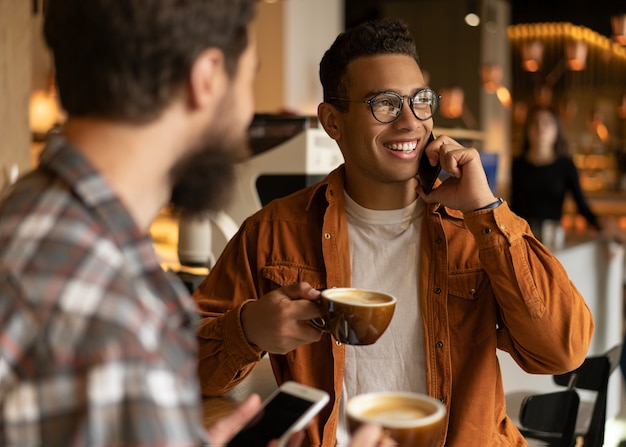 Portrait d'un homme afro-américain souriant parlant sur téléphone mobile. Amis de hipster boire du café ensemble, communication, rire, utiliser des smartphones, assis dans un café. Concept de pause-café