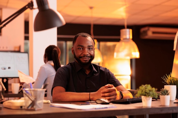 Portrait d'un homme afro-américain travaillant sur ordinateur au bureau pendant le coucher du soleil, en utilisant le réseau de données. Créer une présentation d'entreprise avec des recherches et des informations pour faire un rapport au bureau avec de grandes fenêtres.