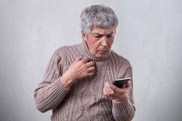 Un portrait d'un homme âgé aux cheveux gris ayant une expression choquée tout en regardant son smartphone lire un message