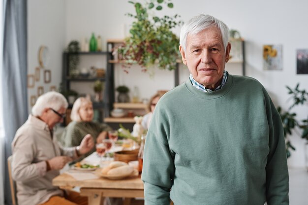 Portrait d'un homme âgé aux cheveux gris regardant la caméra avec ses amis assis à table à manger en arrière-plan