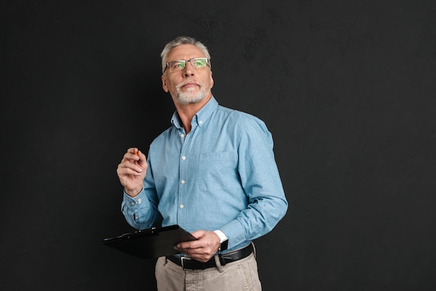 Portrait d'homme d'âge moyen des années 60 avec les cheveux gris et la barbe regardant vers le haut tout en maintenant le presse-papiers avec des documents, isolé sur mur noir