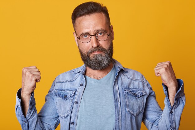 Portrait d'un homme d'âge moyen portant une veste en jean et des lunettes montrant les muscles. Isolé sur mur jaune