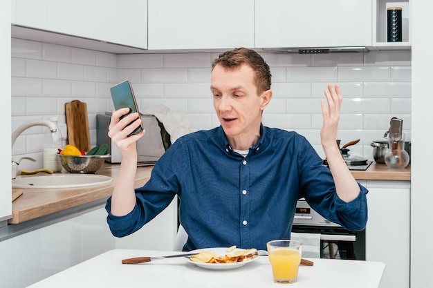 Portrait d'un homme d'âge moyen surpris avec de courts cheveux roux en chemise bleue regardant un téléphone portable avec le bras levé