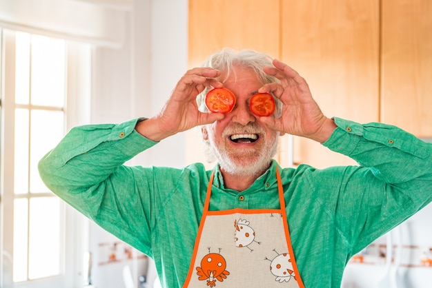 Portrait d'un homme âgé ou mûr riant et souriant en regardant la caméra en train de cuisiner et avec deux tomates sur les yeux faisant une grimace