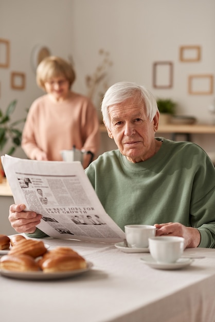 Portrait d'un homme âgé prenant son petit-déjeuner et lisant le journal à table avec sa femme en arrière-plan