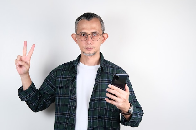 Portrait d'un homme asiatique adulte avec une expression sérieuse montrant un signe de deux doigts et tenant son téléphone