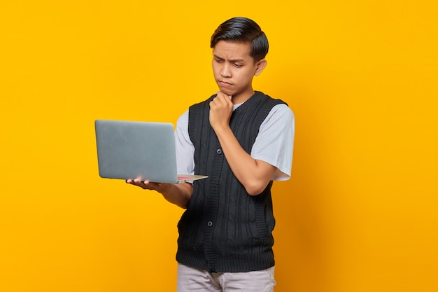 Portrait d'un homme asiatique confiant tenant un ordinateur portable et regardant le courrier électronique entrant sur fond jaune