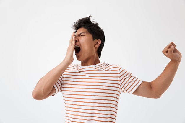 Portrait d'un homme asiatique endormi aux cheveux bruns en t-shirt décontracté bâillant et couvrant la bouche avec le poing en raison de l'insomnie ou de la fatigue, isolé